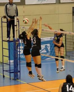 female spiking the volleyball with two blockers, with ref looking on