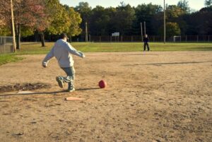 a guy in jeans kicking a dodgeball at home plate on a baseball diamond