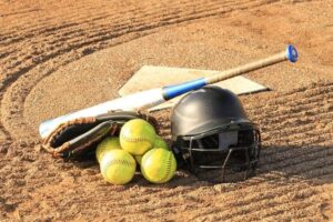 a softball bat leaning on a helmet and a pile of 4 softballs, with home plate in the background