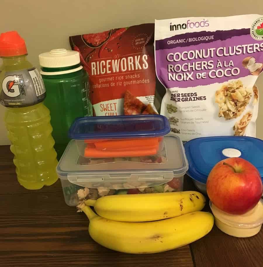 healthy lunch, including bananas, apple, carrot sticks, gatorage, water bottle, rice crackers, and coconut chips