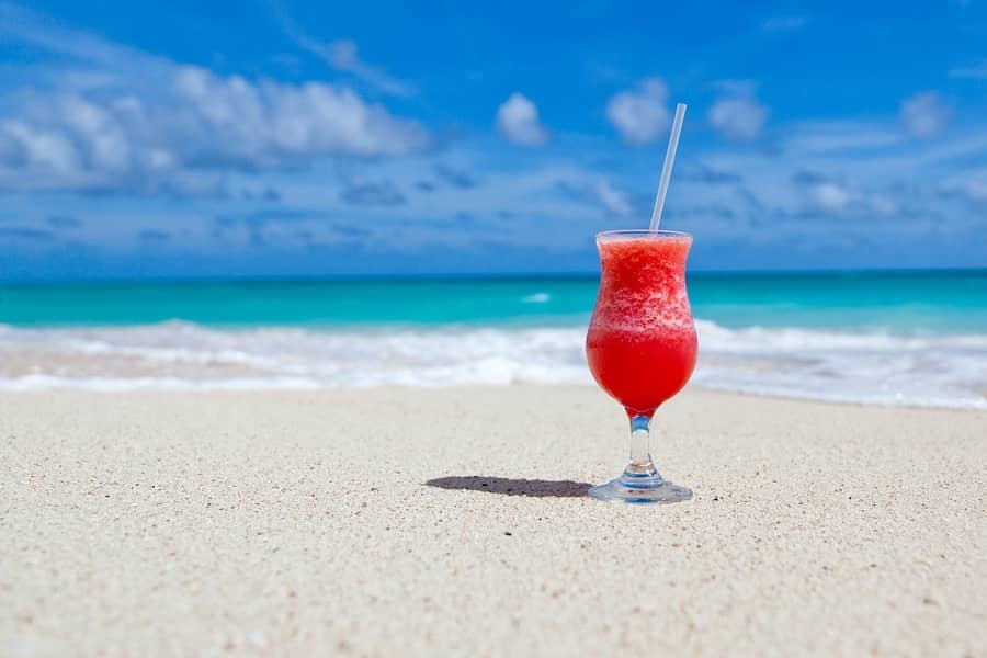 pink cocktail sitting on a sandy beach with blue sky and turquoise water in background